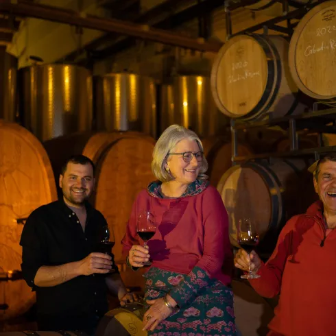 Drei Personen stehen in einem Weinkeller und halten Gläser mit Wein. Im Hintergrund sind große Holzfässer sichtbar.