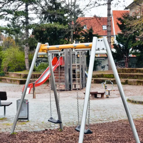 Ein Spielplatz mit einer Schaukel und einer Rutsche. Im Hintergrund sind Bäume und Sitzbänke zu sehen.