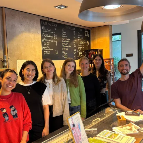 Eine Gruppe von fünf Frauen und einem Mann steht in einem Café hinter einer Theke. Auf der Wand sind Menütafeln sichtbar und die Atmosphäre wirkt freundlich und einladend.