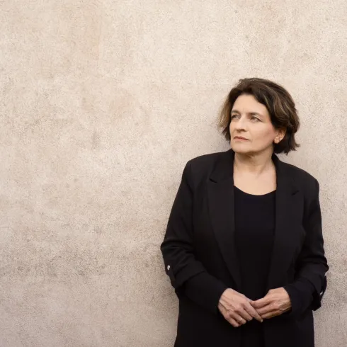 Eine Frau steht an einer grauen Wand und blickt nachdenklich in die Ferne. Sie trägt einen schwarzen Blazer und hat kurze, lockige Haare.
