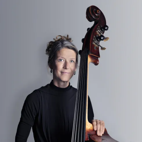 Eine Musikerin steht mit einem Kontrabass vor einem einfarbigen Hintergrund. Sie lächelt und trägt ein schwarzes Oberteil.