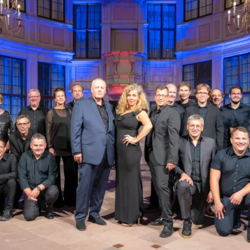 Eine große Gruppe von Menschen steht in einem eleganten Raum mit buntem Licht. Die Männer tragen schwarze Kleidung, während eine Frau in einem eleganten Kleid im Vordergrund posiert.