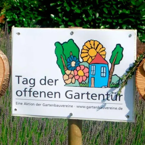 Ein Schild mit der Beschriftung "Tag der offenen Gartentür" steht zwischen zwei Holzgesichtern, die freundlich aussehen. Im Hintergrund sind bunte Blumen und Pflanzen zu sehen.