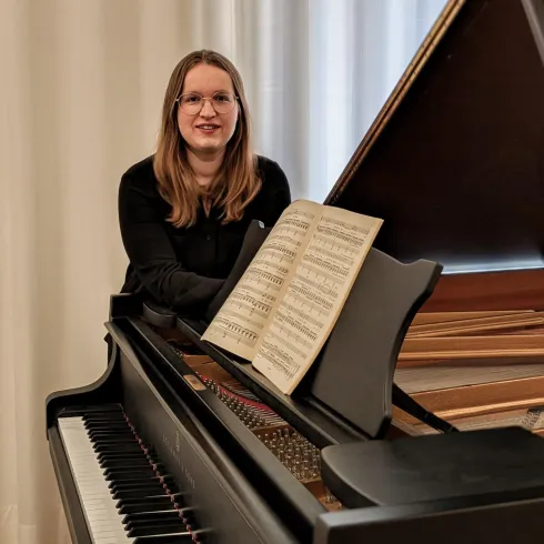 Eine Person sitzt an einem Klavier mit einem aufgeschlagenen Notenblatt. Im Hintergrund sind helle Vorhänge zu sehen.