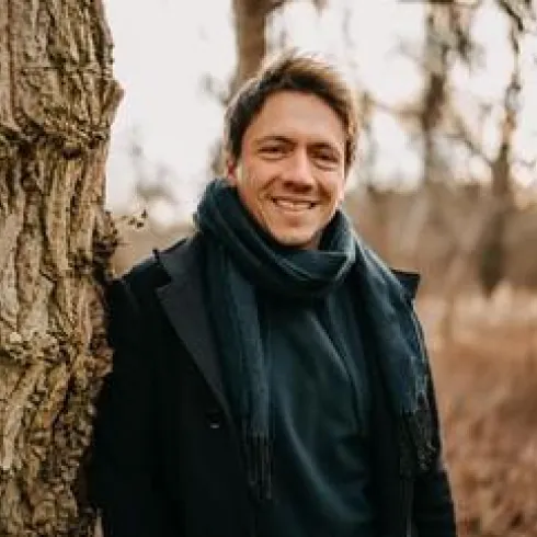 Ein lächelnder Mann steht an einem Baum in einer natürlichen Umgebung. Er trägt einen schwarzen Mantel und einen Schal.