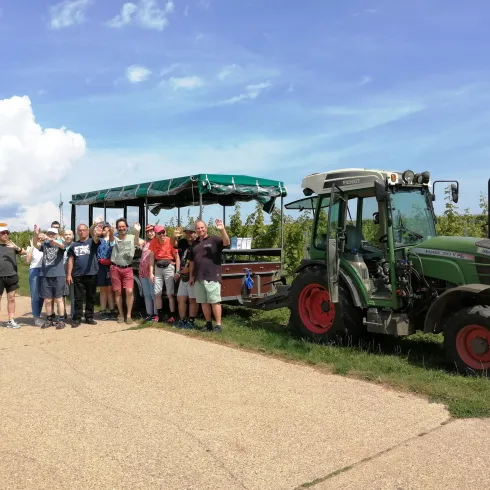 Eine Gruppe von Menschen steht neben einem Traktor und einem Weinfasswagen auf einem Feld. Der Himmel ist blau mit einigen Wolken.