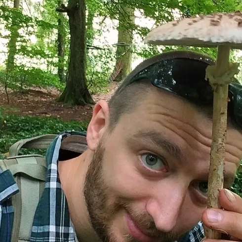 Ein Mann hält einen großen Pilz in einem Wald. Im Hintergrund sind Bäume und grünes Blattwerk zu sehen.