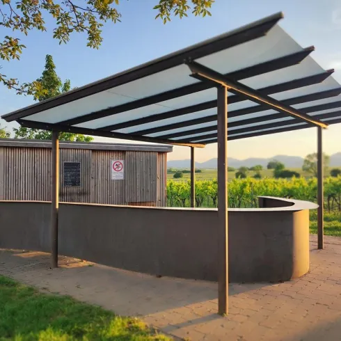 Ein moderner Unterstand mit einem abgedämpften Dach steht inmitten von Weinbergen. Im Hintergrund sind sanfte Hügel und Bäume zu sehen.