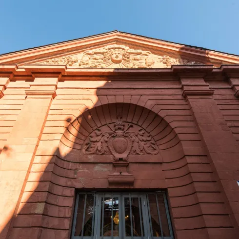 Ein historisches Gebäude mit einer markanten roten Fassade. Das Detail über dem Eingang zeigt kunstvolle Ornamente.