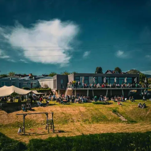 Eine lebhafte Veranstaltung im Freien mit vielen Menschen, die auf einer Terrasse stehen. Im Vordergrund sind eine Hängematte und ein Spielplatz sichtbar.