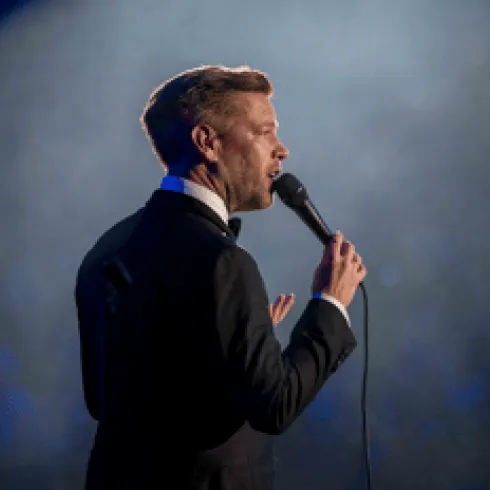 Ein Mann in einem Anzug steht auf der Bühne und spricht in ein Mikrofon. Im Hintergrund ist ein sanftes Licht zu sehen.