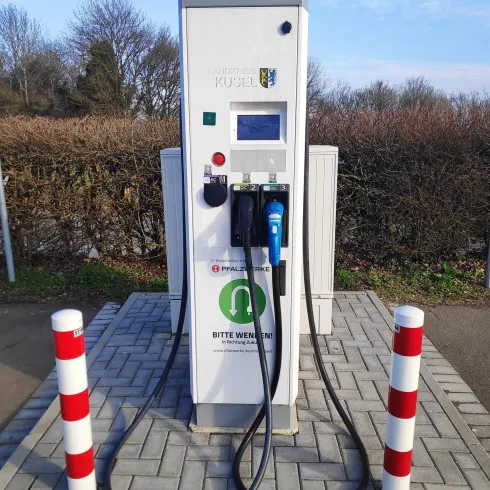 Eine Elektroladestation steht auf einem gepflasterten Platz. Sie ist mit Kabeln und umgeben von Absperrpfosten.