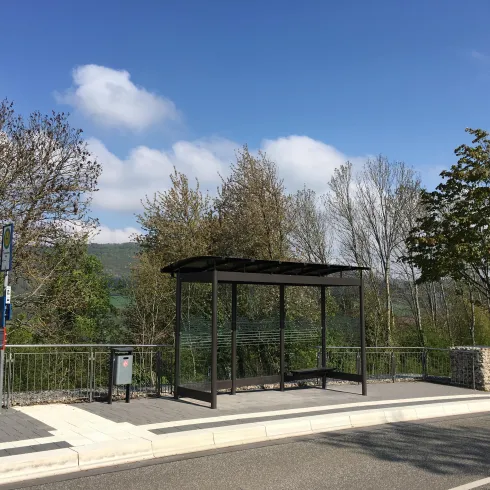 Eine Bushaltestelle in ländlicher Umgebung mit wenigen Bäumen und blauem Himmel. Die Haltestelle hat ein modernes, einfaches Design.