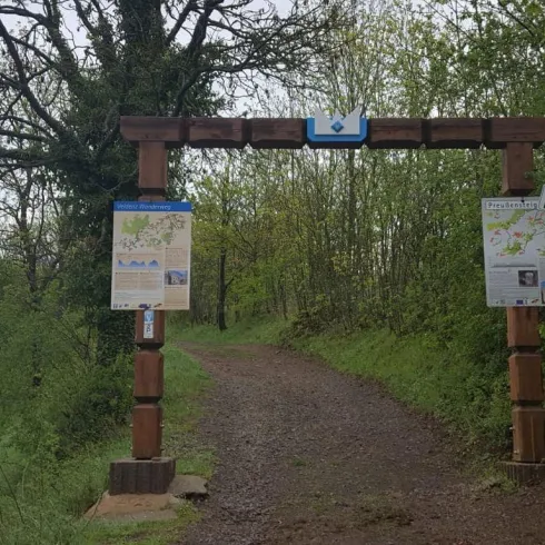 Ein Eingang zu einem Wanderweg mit einem hölzernen Tor und Informationsschildern. Umgeben von Bäumen und einer grünen Landschaft.