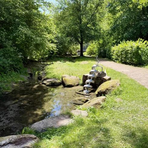 Ein idyllischer Weg entlang eines kleinen Bachs. Grüne Bäume und Sträucher umgeben den Bereich, während ein Wasserspiel aus Steinen im Vordergrund zu sehen ist.