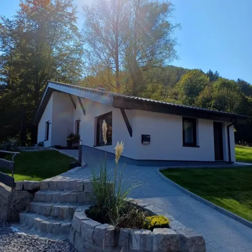 Ein modernes Haus mit hellem Außenanstrich steht umgeben von Bäumen und gepflegtem Rasen. Der Weg zum Haus ist mit Steinen und Pflanzen dekoriert.