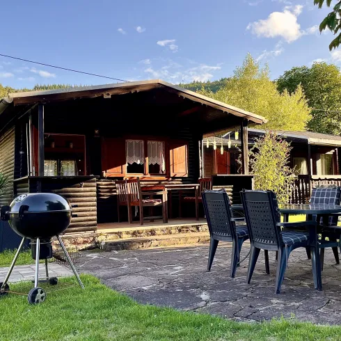 Ein gemütliches Ferienhaus im Grünen mit einem Grill und einem Esstisch auf der Terrasse. Umgeben von Bäumen und einer schönen Landschaft.