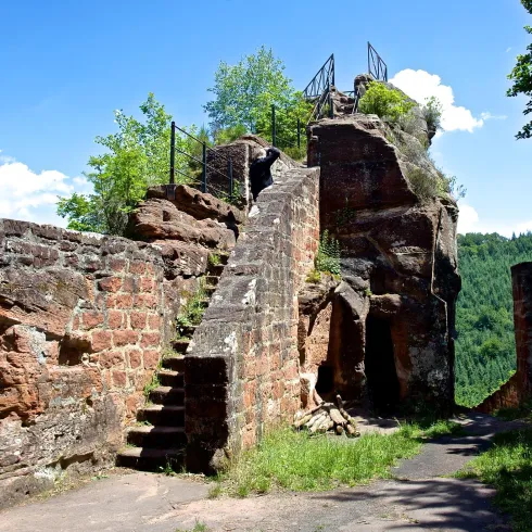 Eine alte Ruine mit steinernen Wänden und einer Treppe, die zu einem Aussichtspunkt führt. Im Hintergrund sind grüne Wälder und ein blauer Himmel sichtbar.