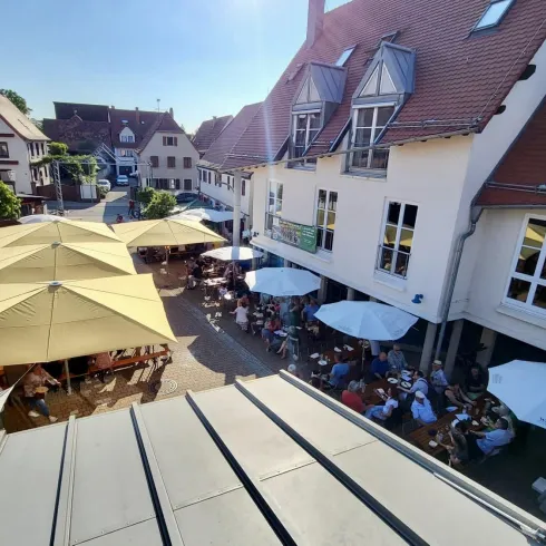 Eine lebhafte Außenterrasse mit vielen Gästen unter Sonnenschirmen. Umgeben von charmanten Gebäuden in einer sonnigen Umgebung.