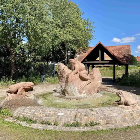 Eine kunstvolle Statue eines Frosches, die aus Stein besteht, steht im Zentrum eines Brunnens. Um den Brunnen herum sind weitere Frösche angeordnet, umgeben von einer grünen Landschaft.