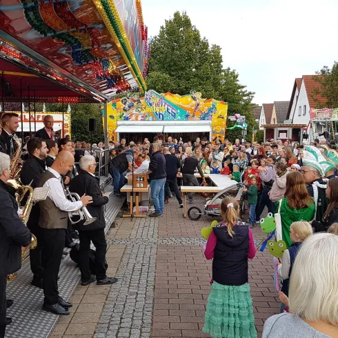 Eine lebhafte Menschenmenge genießt ein Fest, während eine Band auf einer Bühne Musik spielt. Bunte Attraktionen und festliche Stimmung prägen die Szene.