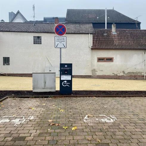 Ein leerer Parkplatz mit einem Parkscheinautomaten und einem Schild, das das Parken verbietet. Im Hintergrund sind verschiedene Gebäude zu sehen.