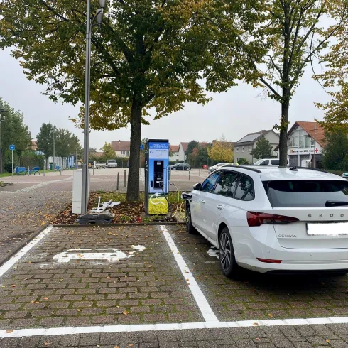 Ein parkendes weißes Auto steht auf einem Parkplatz mit Ladegerät. Im Hintergrund sind Bäume und ein ruhiger Ort zu sehen.