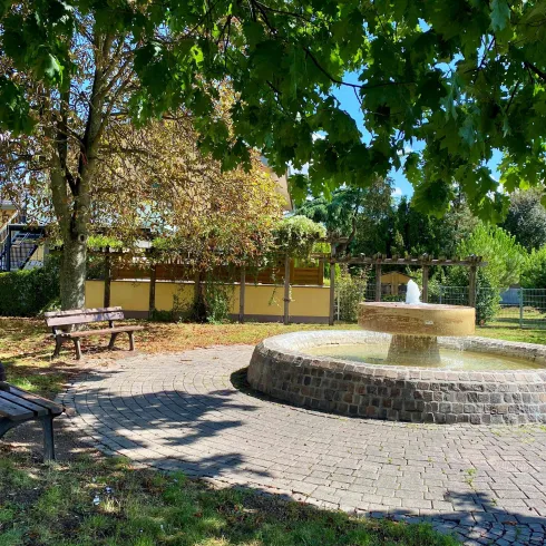 Ein schöner Park mit einem Brunnen in der Mitte. Bänke laden zum Entspannen ein, umgeben von Bäumen und Grünflächen.