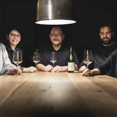 Eine Gruppe von fünf Personen sitzt an einem Holztisch. Sie lächeln und halten Weingläser, während eine Flasche Wein in der Mitte steht.