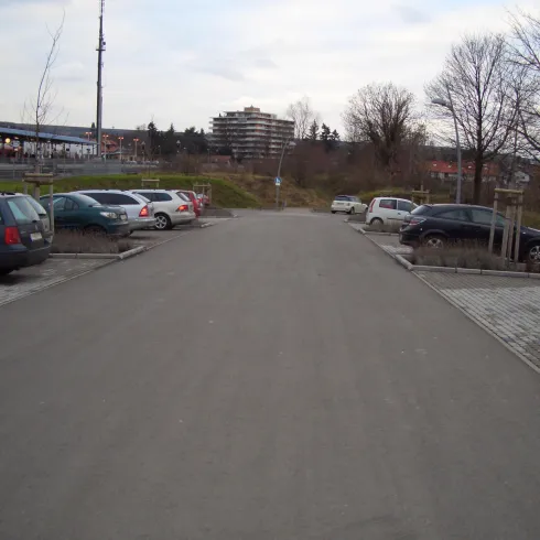 Ein Parkplatz mit mehreren Autos und einer leeren Fahrbahn. Im Hintergrund sind Gebäude und Bäume sichtbar.
