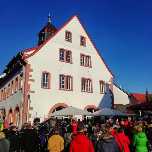 Eine Gruppe von Menschen versammelt sich vor einem historischen Gebäude mit einer roten und weißen Fassade. Der Himmel ist klar und blau, was eine schöne Atmosphäre schafft.