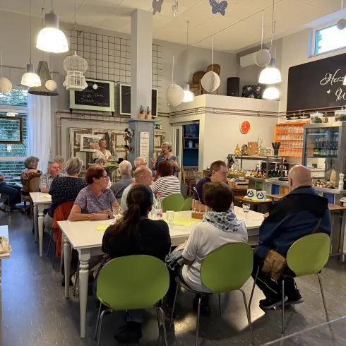 Ein einladendes Café mit mehreren Tischen, an denen Gäste sitzen und sich unterhalten. Helle Wände und dekorative Elemente schaffen eine gemütliche Atmosphäre.