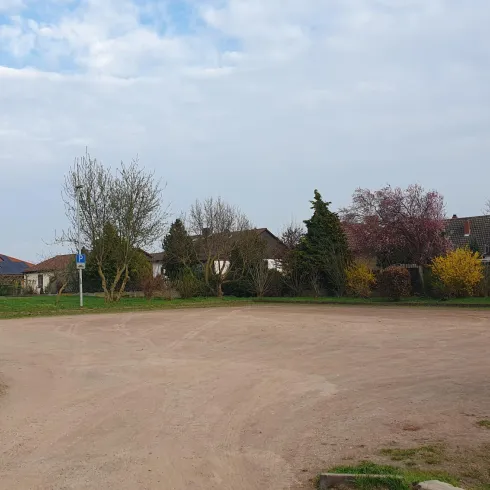 Ein Parkplatz mit ein paar Abfallcontainern und einigen Bäumen. Im Hintergrund sind mehrere Wohnhäuser zu sehen.