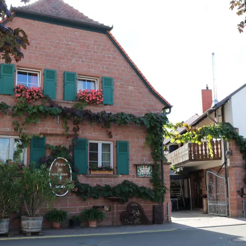 Ein charmantes Haus mit roten Backsteinen und grünen Fensterläden, umgeben von Pflanzen. Das Gebäude hat einen einladenden Eingang mit Blumen.