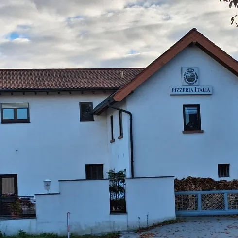 Ein weißes Gebäude mit einem braunen Dach, das die Pizzeria Italia beherbergt. Der Eingang und der Gartenbereich sind gut sichtbar.