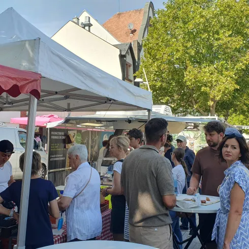 Ein lebhafter Markt mit mehreren Ständen und vielen Menschen. Im Hintergrund sind Bäume und Gebäude zu sehen.