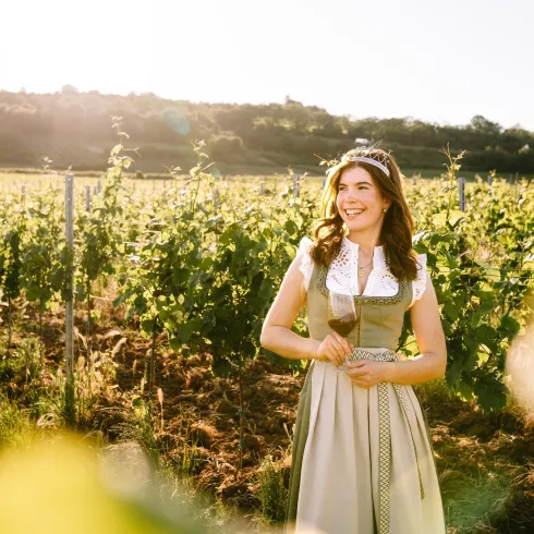 Eine fröhliche Frau steht in einem Weinberg. Die Sonne scheint und die Trauben wachsen in der Nähe.