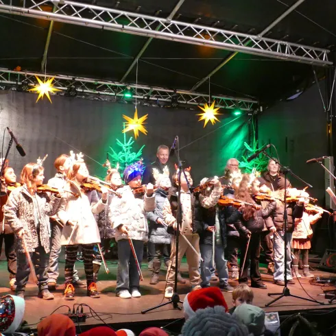 Eine Gruppe von Kindern und Erwachsenen spielt auf der Bühne Weihnachtsmusik. Die Szene ist festlich mit bunten Lichtern und Sternen dekoriert.