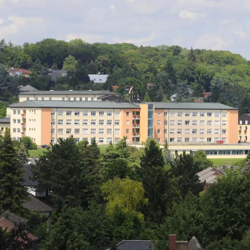 Ein modernes Gebäude, umgeben von Bäumen und Bergen. Der Himmel ist teilweise bewölkt.