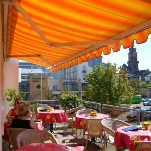 Eine sonnige Terrasse mit bunten Tischen und Stühlen. Im Hintergrund ist eine Stadtansicht mit einem alten Gebäude und Bäumen zu sehen.