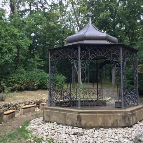 Ein schöner Pavillon aus Metall in einem grünen Park. Umgeben von Bäumen und einem kleinen Steinrand.