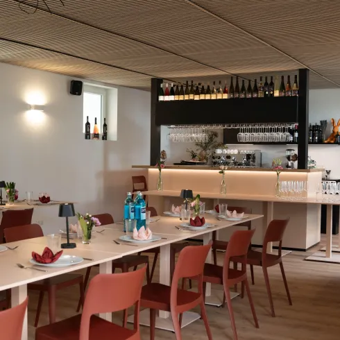 Ein modernes Restaurant mit roten Stühlen und hellen Tischen. Die Bar im Hintergrund ist dekoriert und es gibt Gläser und Flaschen auf dem Regal.