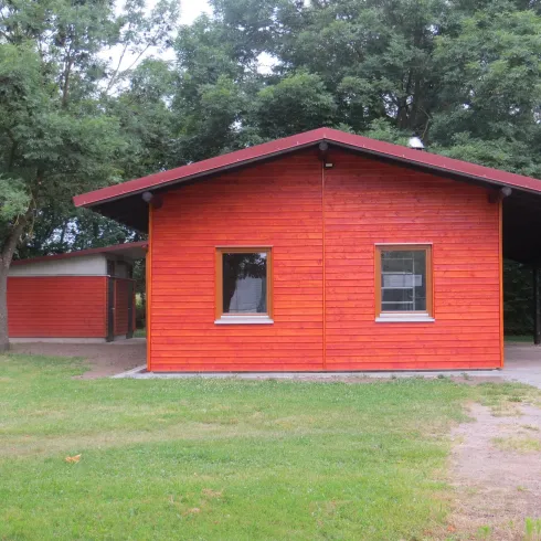 Ein rotes Holzhaus steht in einem grünen Garten. Bäume umgeben das Gebäude und schaffen eine ruhige Atmosphäre.
