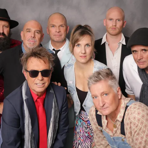 Eine Gruppe von neun Personen posiert gemeinsam für ein Foto. Die Menschen tragen verschiedene stilvolle Outfits und lächeln.
