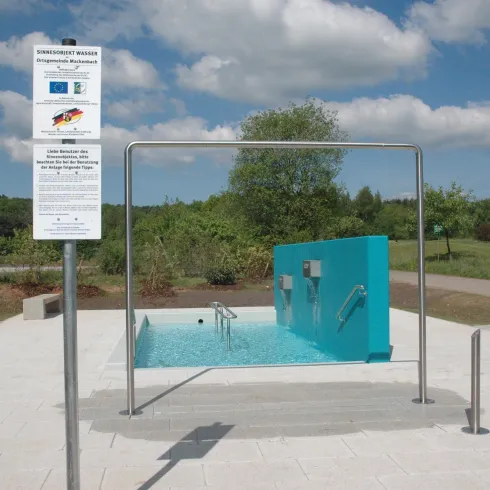 Ein moderner Wasserfilter mit blauer Wand, umgeben von einer grünen Landschaft. Die Anlage steht auf einem hellen Platz unter einem klaren, blauen Himmel.