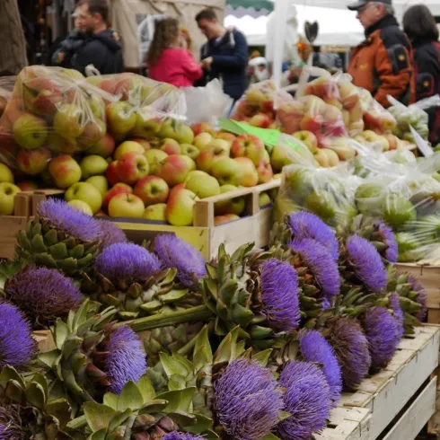 Ein lebhafter Markt mit frischen Äpfeln und bunten Artischocken. Menschen stehen im Hintergrund und genießen das bunte Angebot.