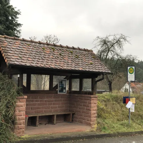 Eine Bushaltestelle aus Holz mit einem schrägen Dach. Im Hintergrund sind Bäume und ein Informationsschild zu sehen.