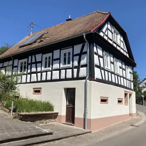 Ein charmantes Fachwerkhaus an einer Ecke, umgeben von grüner Vegetation. Der Himmel ist klar und blau.