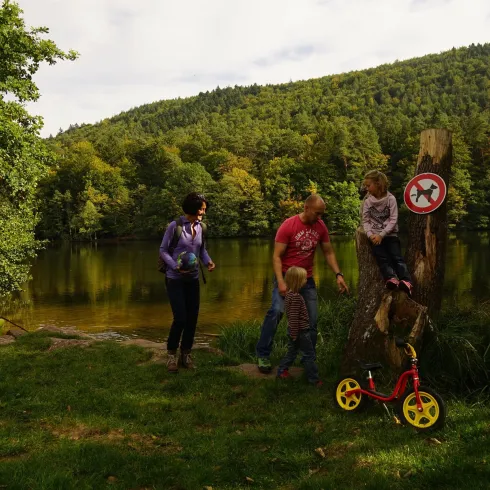 Eine Familie verbringt Zeit am Ufer eines Sees in der Natur. Im Hintergrund sind Bäume und eine ruhige Landschaft zu sehen.