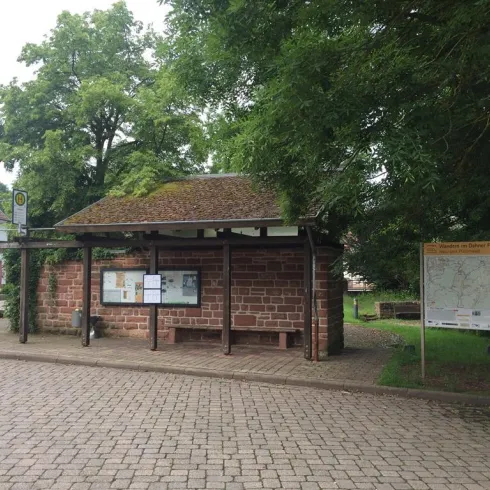 Eine Bushaltestelle aus Stein mit einem begrünten Dach. Im Hintergrund sind Bäume und ein Informationsschild sichtbar.